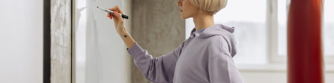 Fit female trainer writes workout plan on a whiteboard in a gym setting. Side view.